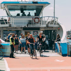 Cyklister och fotgängare går i land från en färja en solig dag, med människor i avslappnad klädsel och en person i en säkerhetsväst.