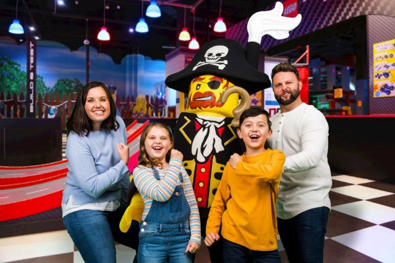 A smiling family poses with a large LEGO pirate mascot in an indoor amusement park, with colorful lights and scenery in the background.