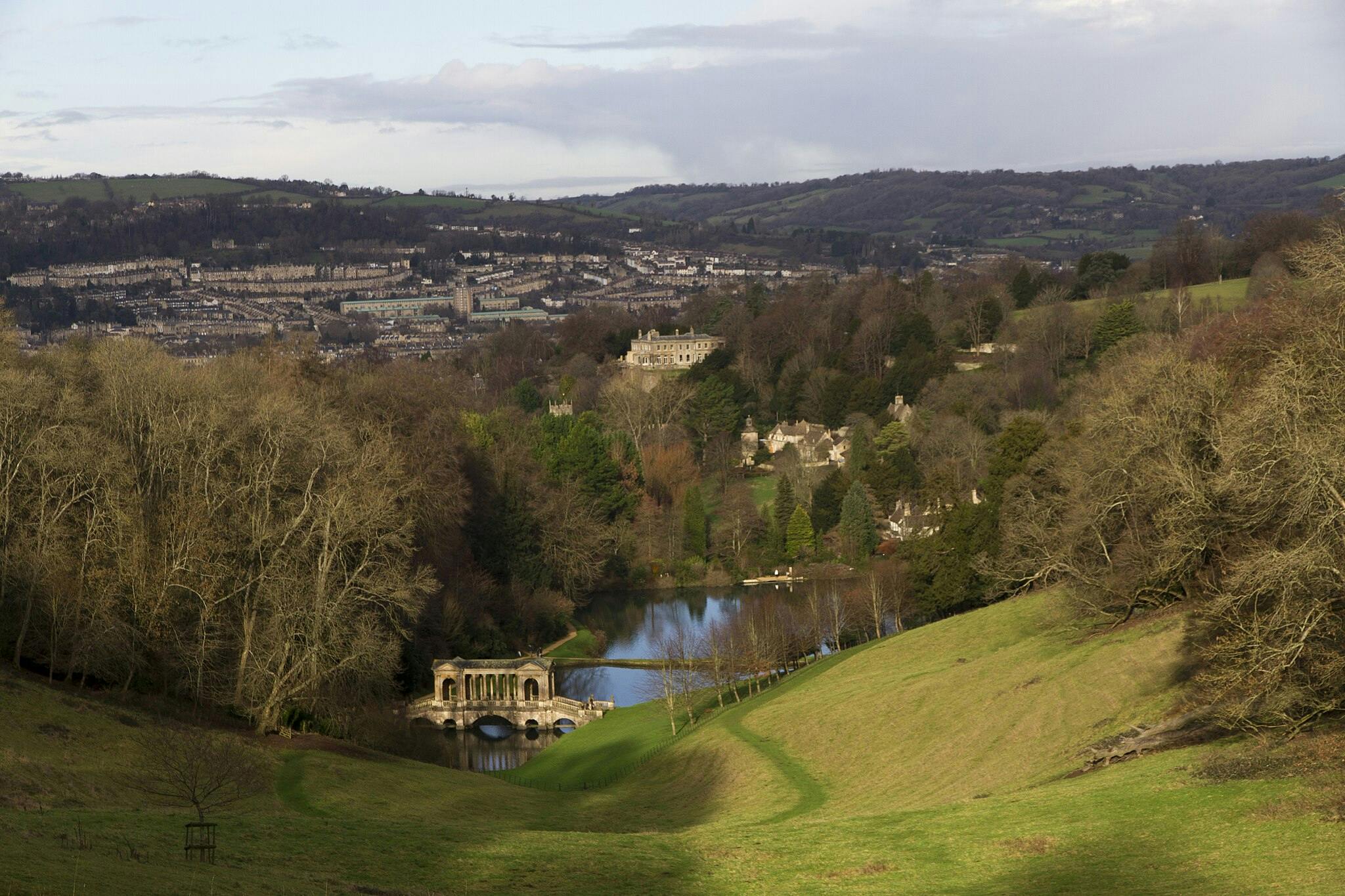 시골 풍경은 시냇물 위에 있는 유서 깊은 다리, 구불구불한 푸른 언덕, 맨발의 나무, 석조 건물이 있는 먼 마을을 특징으로 합니다.