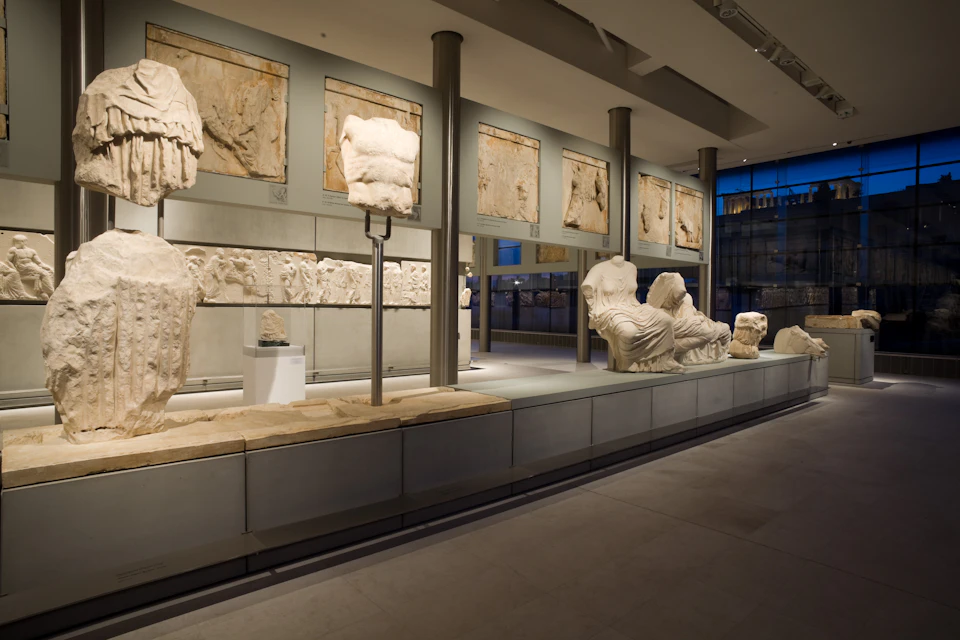 Exhibit of ancient marble sculptures and friezes in a modern museum with large windows and overhead lighting.