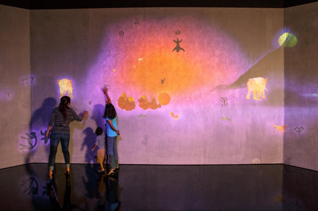 Tres persones interactuen amb una colorida pantalla de paret projectada amb formes d'animals i patrons abstractes.