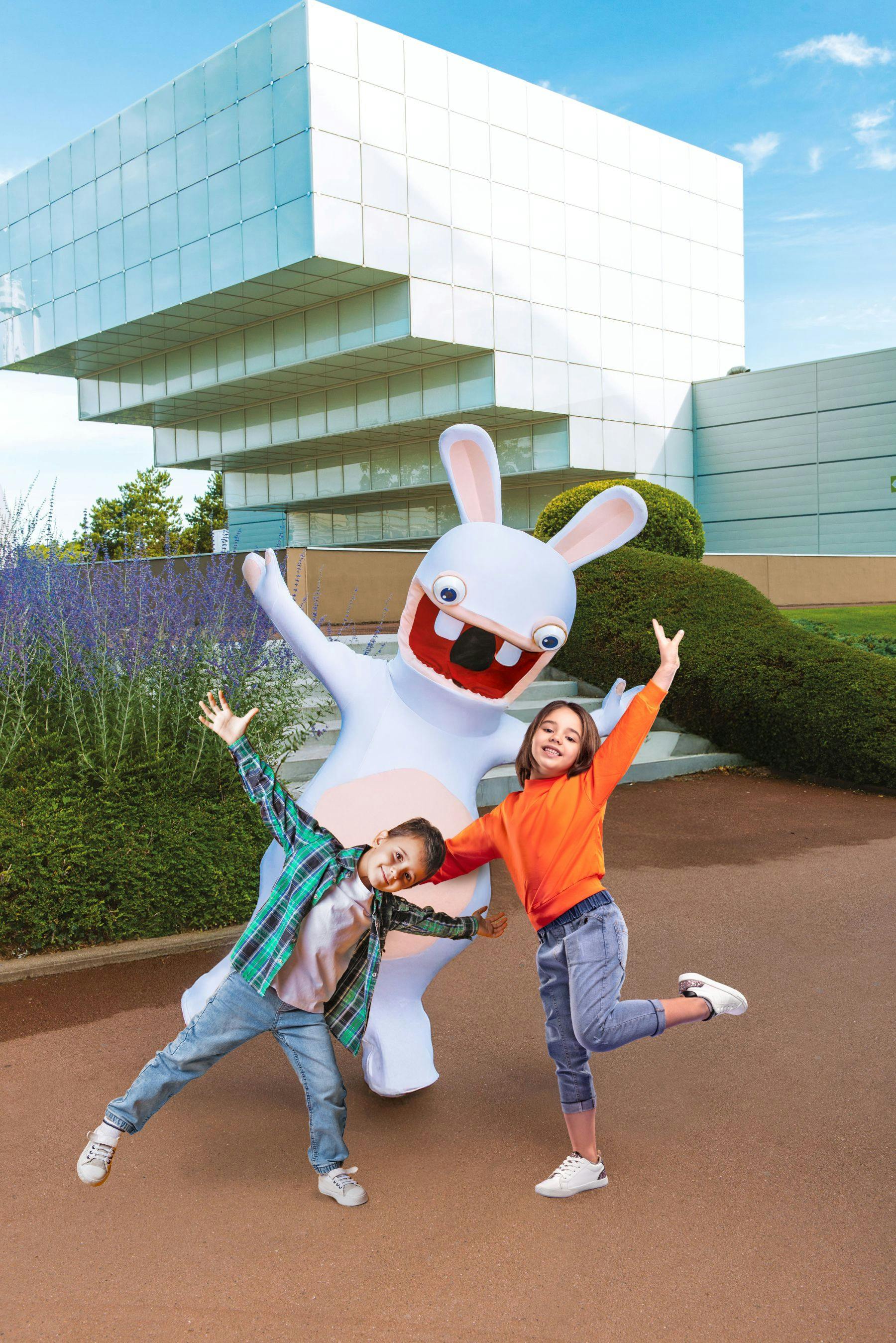 Two children pose playfully with a person in a rabbit costume in front of a modern building and green bushes.