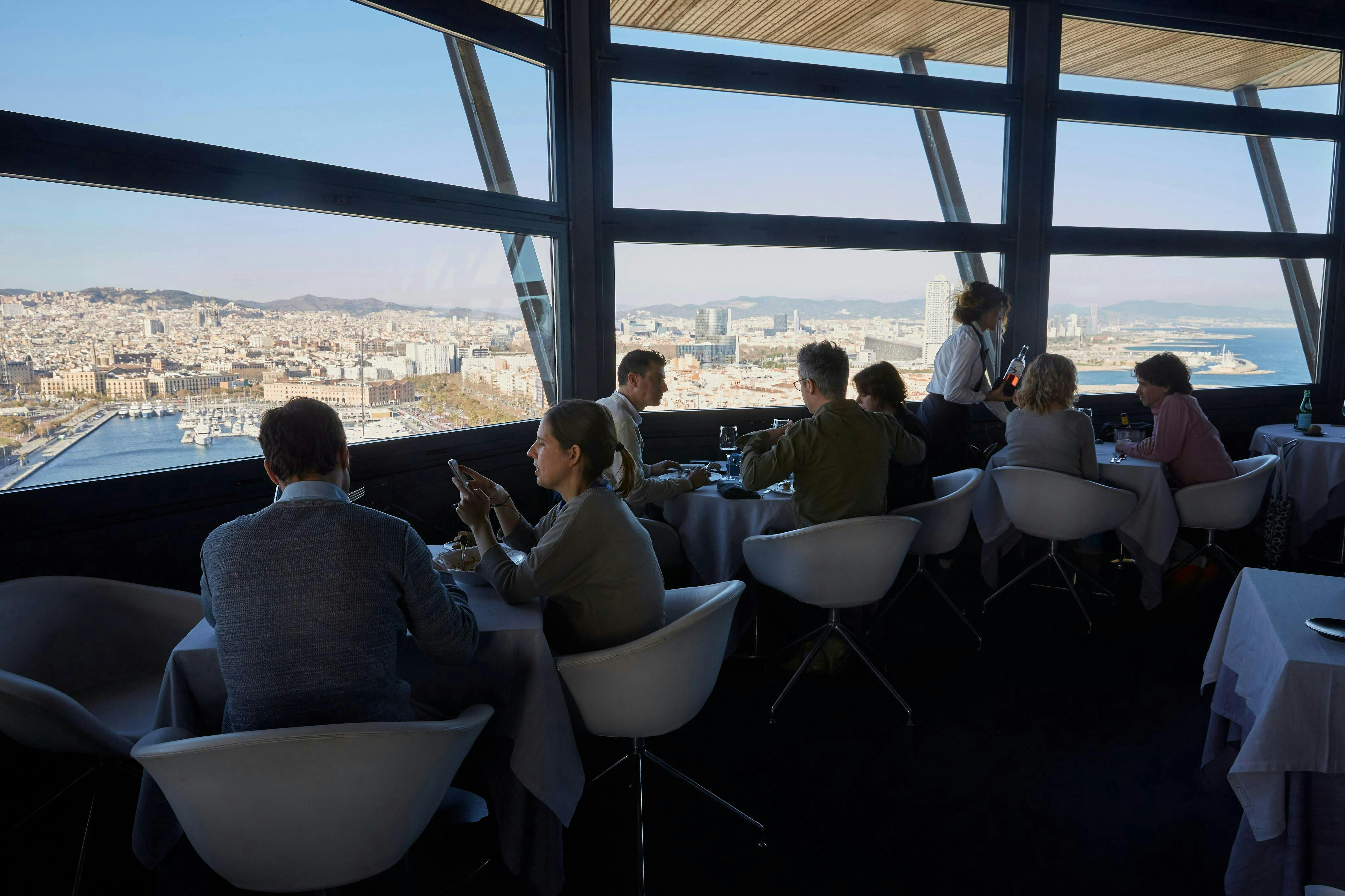 Människor som äter på en restaurang med stora fönster som erbjuder panoramautsikt över en stadsbild och hamn.