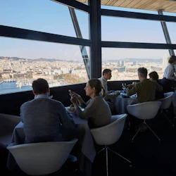 Människor som äter på en restaurang med stora fönster som erbjuder panoramautsikt över en stadsbild och hamn.