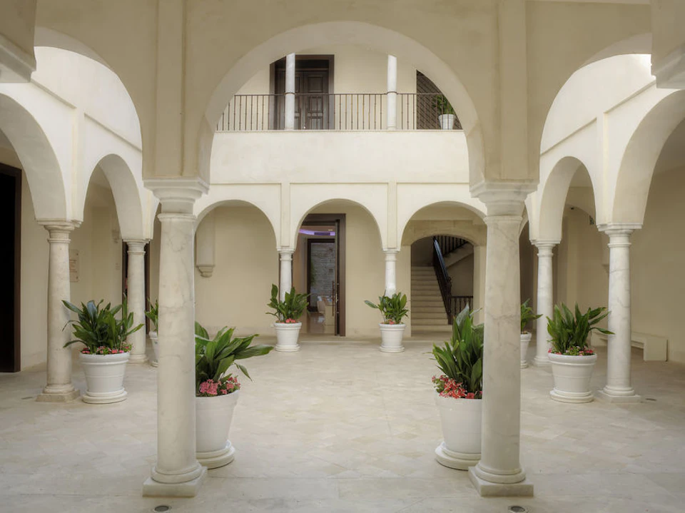 A courtyard with arched columns, potted plants, a staircase, and an upper balcony with doors.