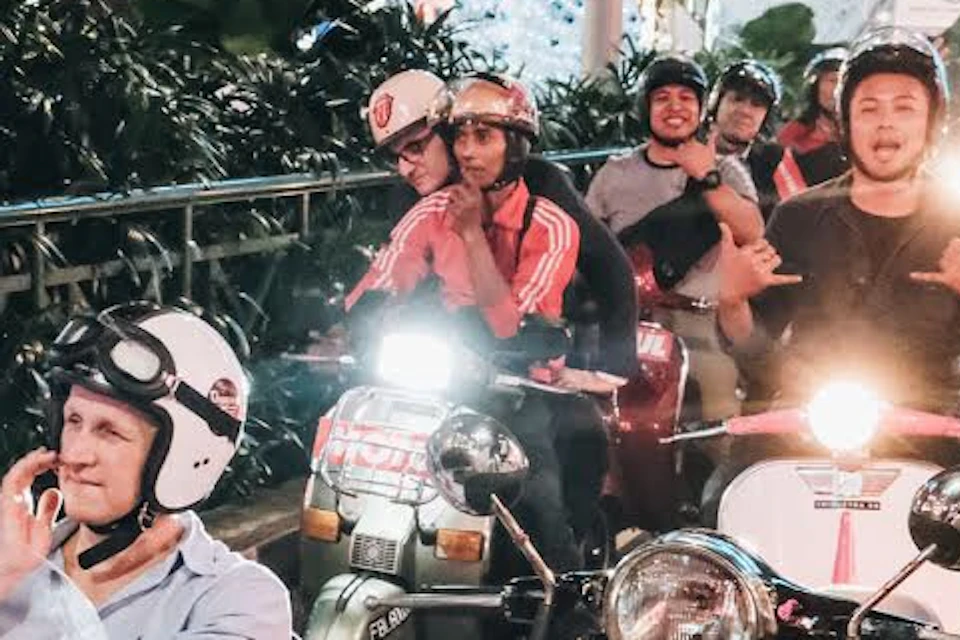 A group of people riding motorcycles and sidecars down a city street at night, waving and smiling.