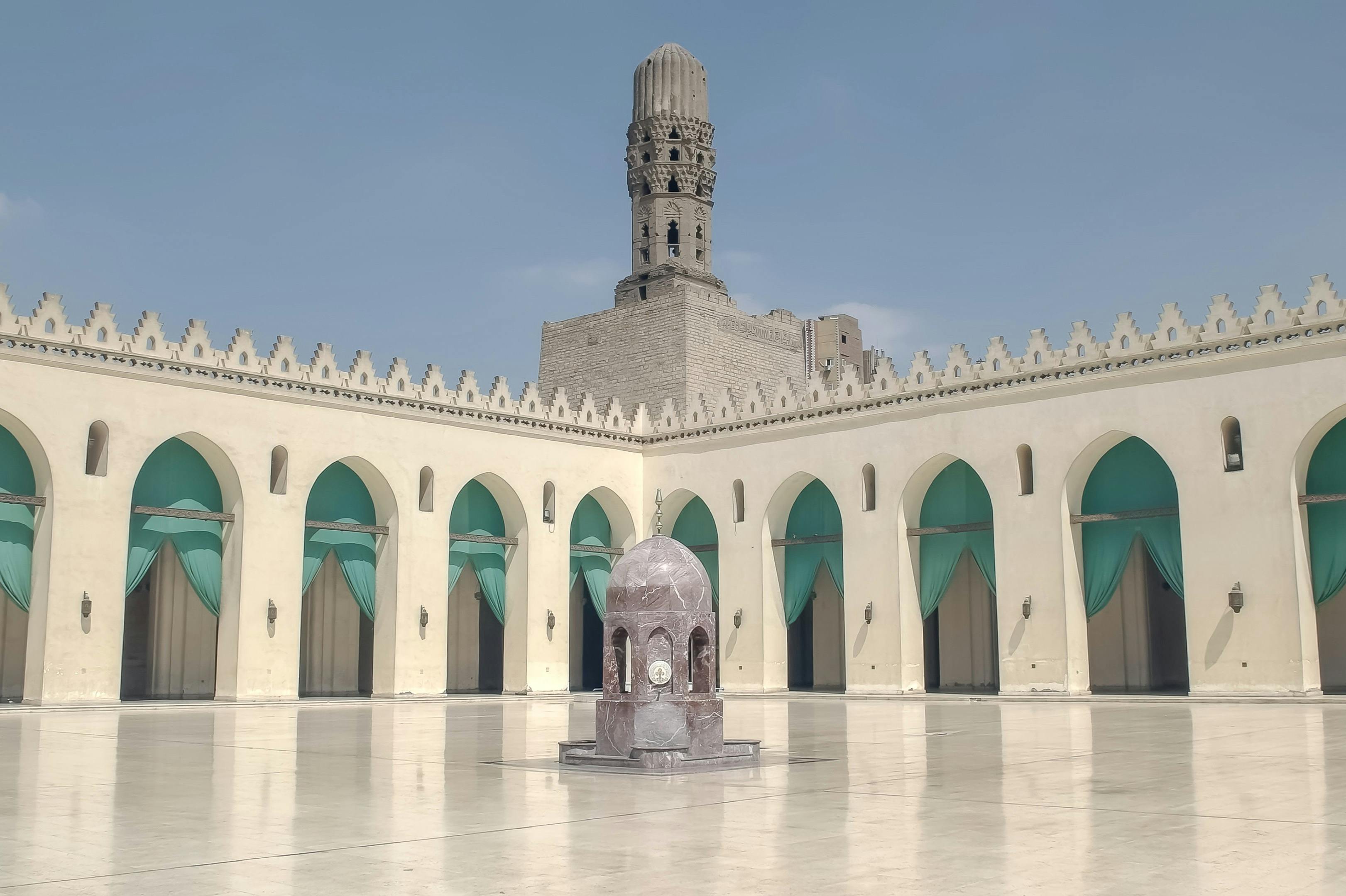 Открытый двор с центральным фонтаном, окруженный арочными дверными проемами, и высоким, богато украшенным минаретом на заднем плане.