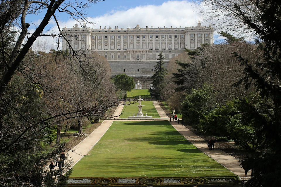 A large, ornate building sits atop a hill, overlooking a manicured garden with a central path and a fountain.