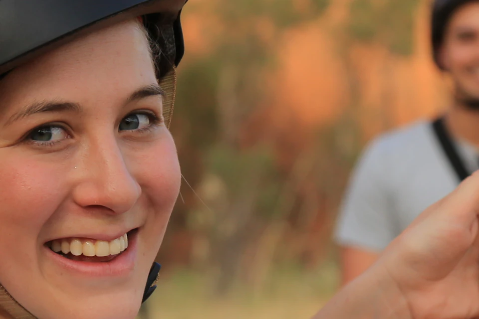 Woman in helmet smiling, holding a smartphone showing a selfie, man standing on a Segway in the background.