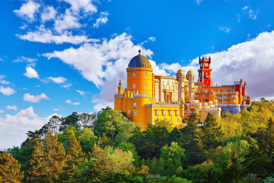 A colorful castle with yellow and red towers surrounded by lush greenery under a bright blue sky with clouds.