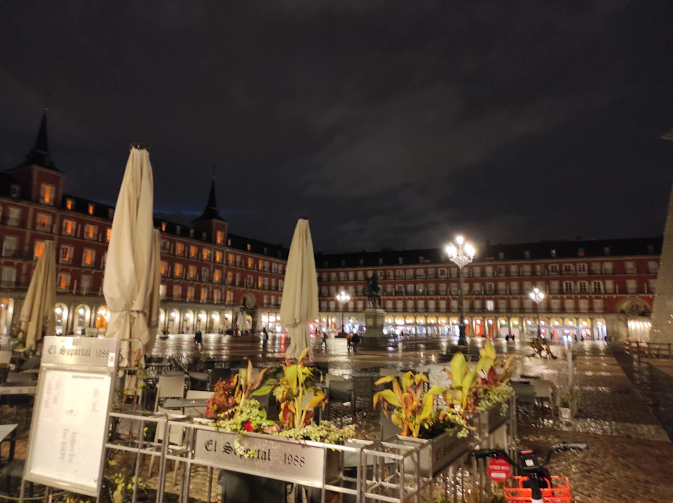 Night view of a square with lit lampposts, closed umbrellas, potted plants, and a statue in the center, with a building in the background.