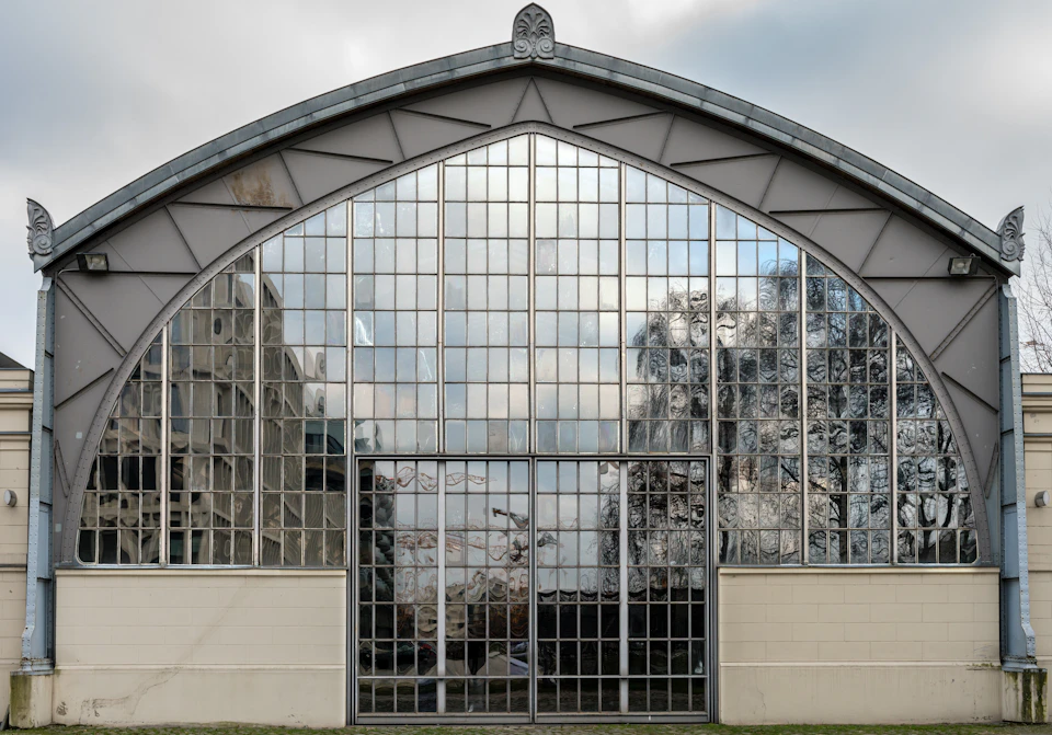 A large arched building entirely covered with reflective glass panels, showing reflections of trees and other structures.