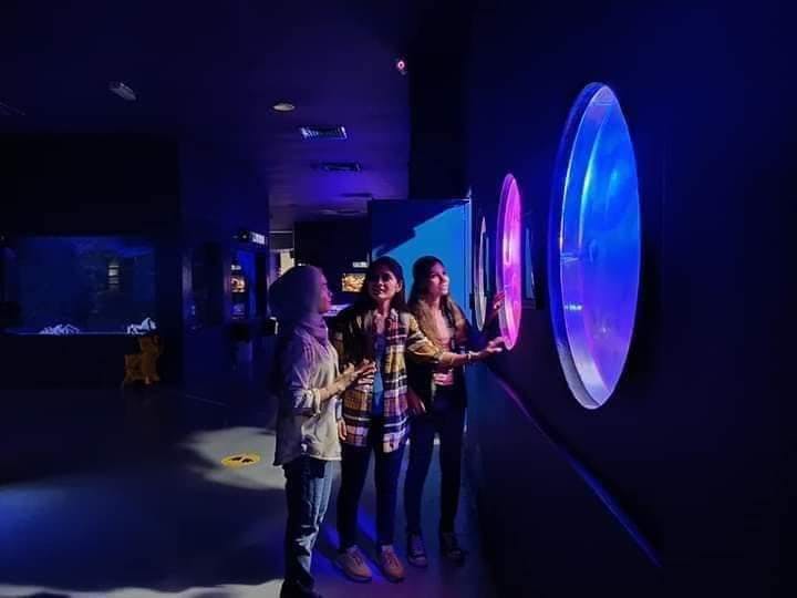 Tre persone interagiscono con display circolari illuminati su una parete scura, in uno spazio espositivo moderno e poco illuminato.