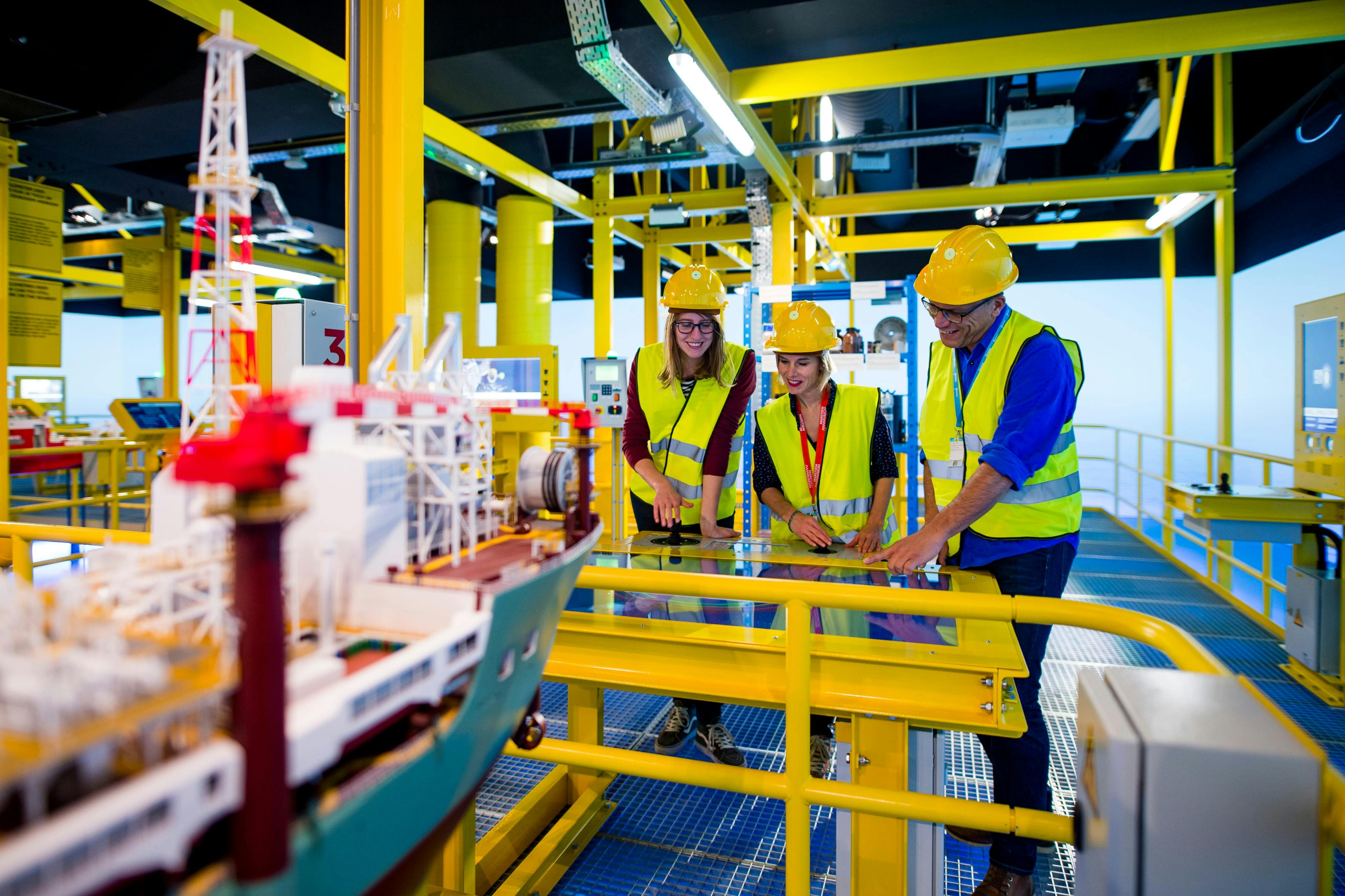三个戴着安全帽和背心的人在工业主题环境中检查一艘模型船和设备。
