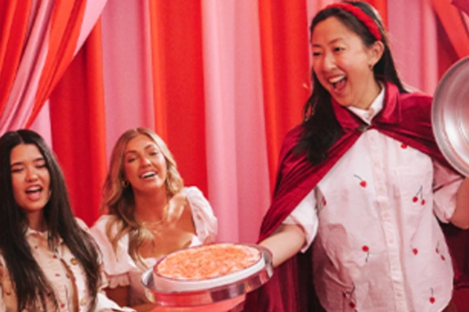 A group of people at a red counter, with a man in a red suit and a woman holding a pie in a colorful, draped room.