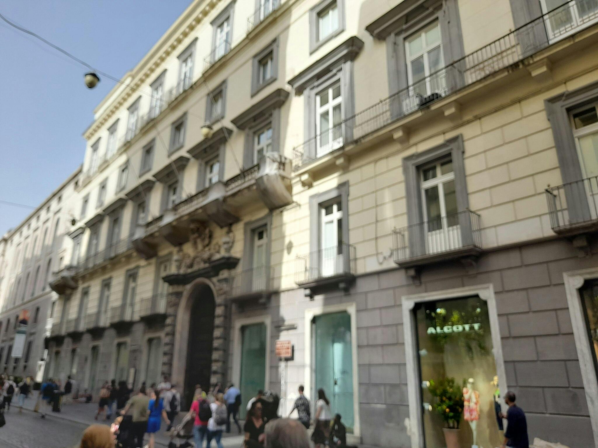 Gade med mennesker, der går foran en historisk bygning med flere etager. En butik med navnet &#34;ALGOTT&#34; er synlig.