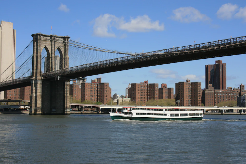 A boat passes under the Brooklyn Bridge with a backdrop of city buildings and a clear, blue sky.
