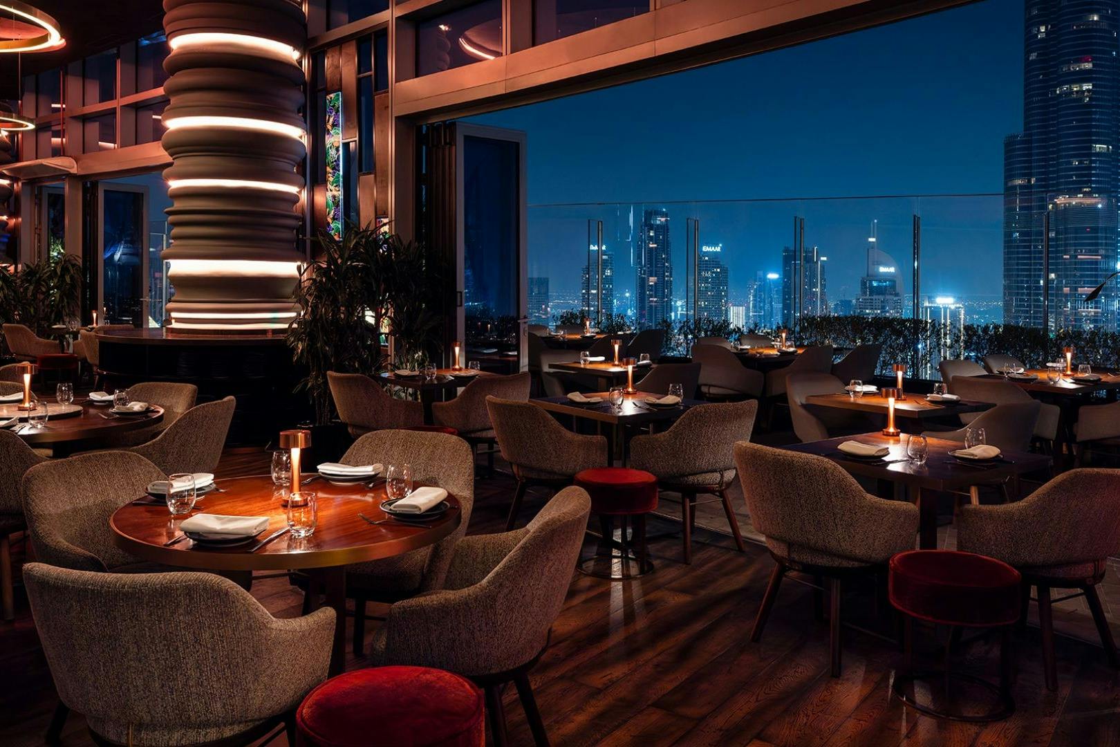 Upscale restaurant with candlelit tables, modern decor, and large windows offering a view of a city skyline at night.