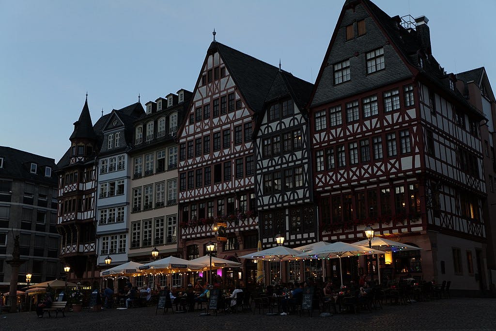 Vista serale di una storica piazza della città con edifici a graticcio e caffè all'aperto, con persone che cenano sotto ombrelloni illuminati.
