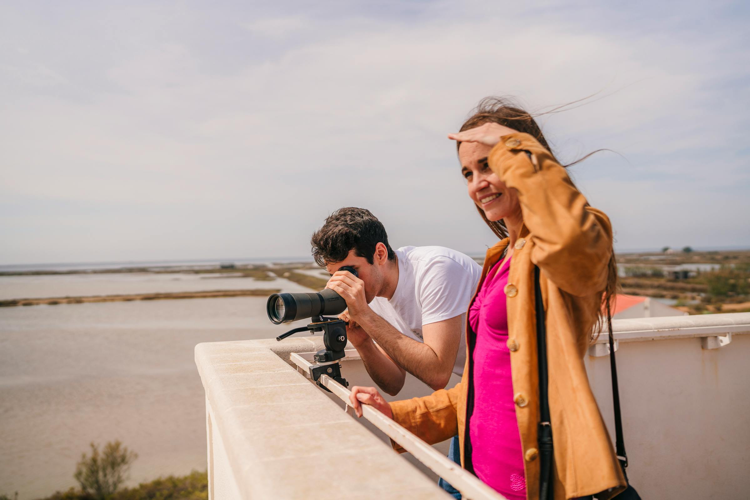 Muž a žena na střeše, muž používá kameru s dlouhým objektivem, zatímco žena stínuje oči rukou a usmívá se.