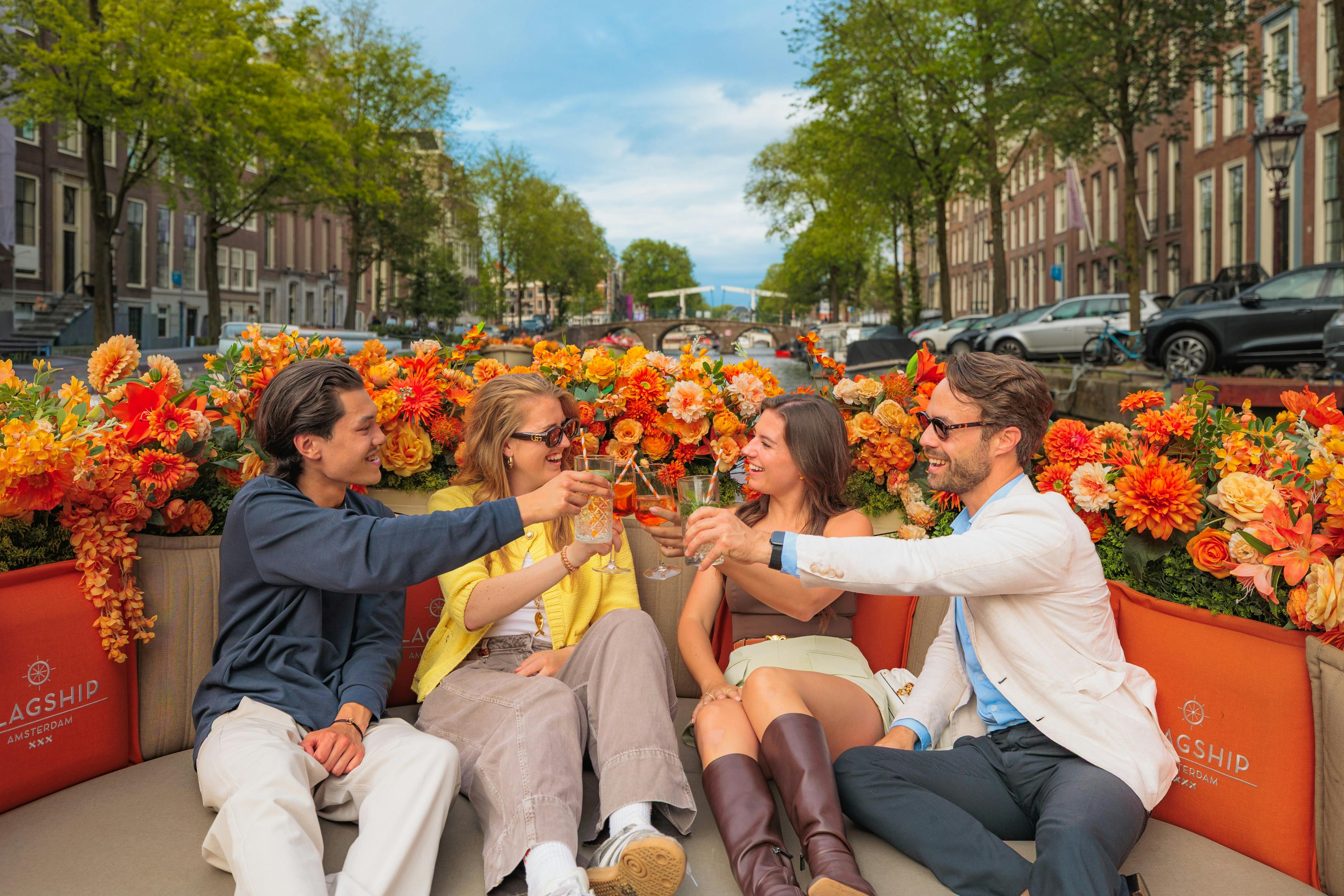 Four people sitting on a boat decorated with orange flowers, smiling and toasting drinks, with a canal and houses in the background.