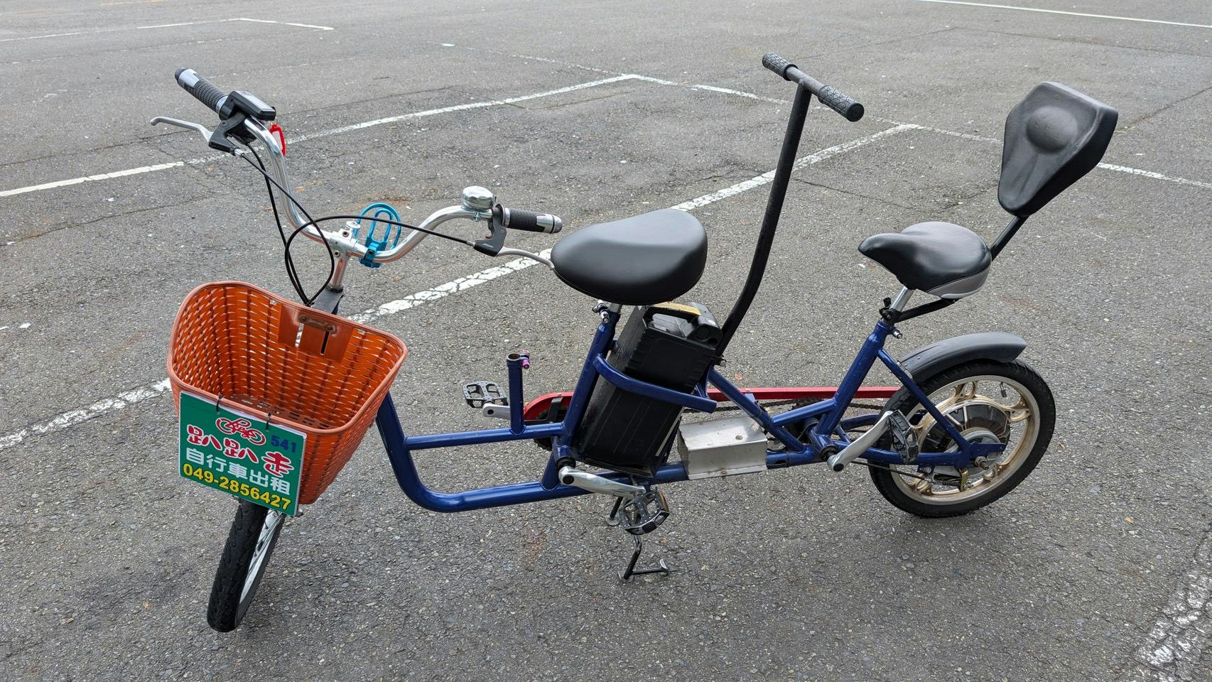 A two-seater bicycle with a blue frame, an orange basket, and an electric battery on a paved surface in a parking area.