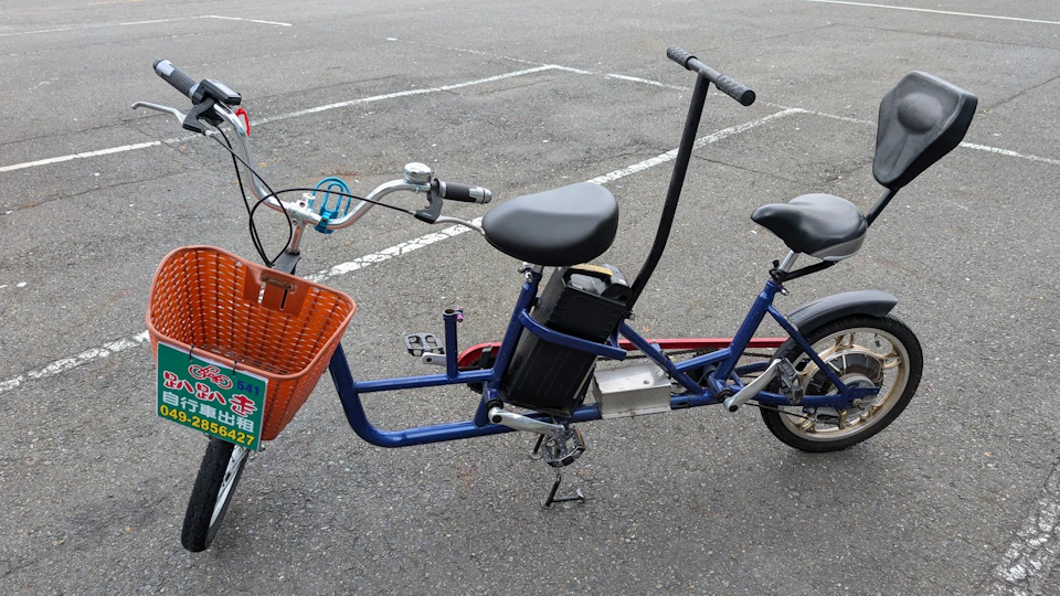 A two-seater bicycle with a blue frame, an orange basket, and an electric battery on a paved surface in a parking area.
