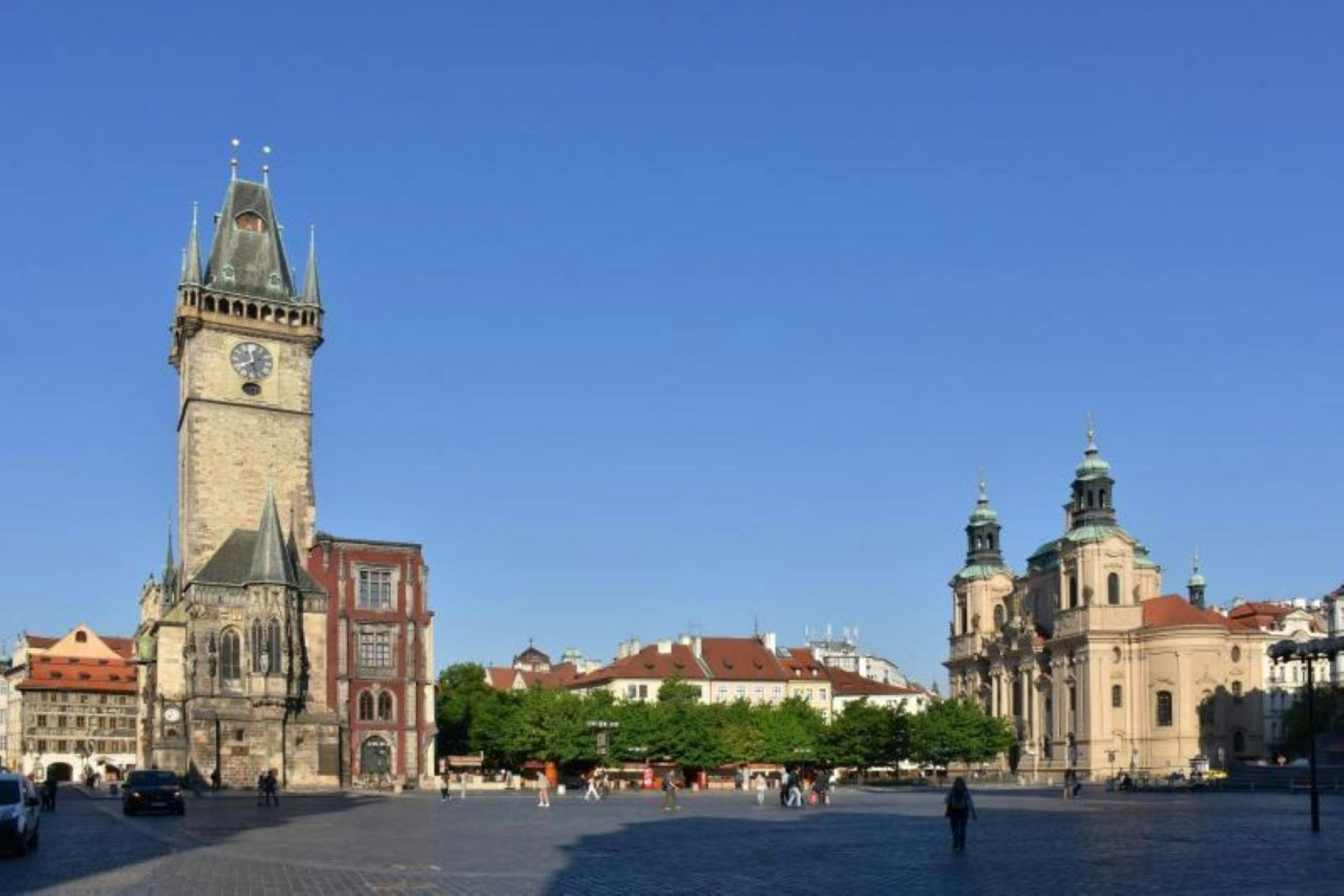A wide square with a historic clock tower on the left, a baroque church on the right, and people walking under a clear blue sky.