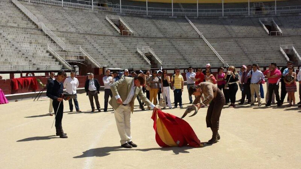 Two people in the foreground engage in a bullfighting demonstration with a red and yellow cape while a crowd watches in an empty arena.