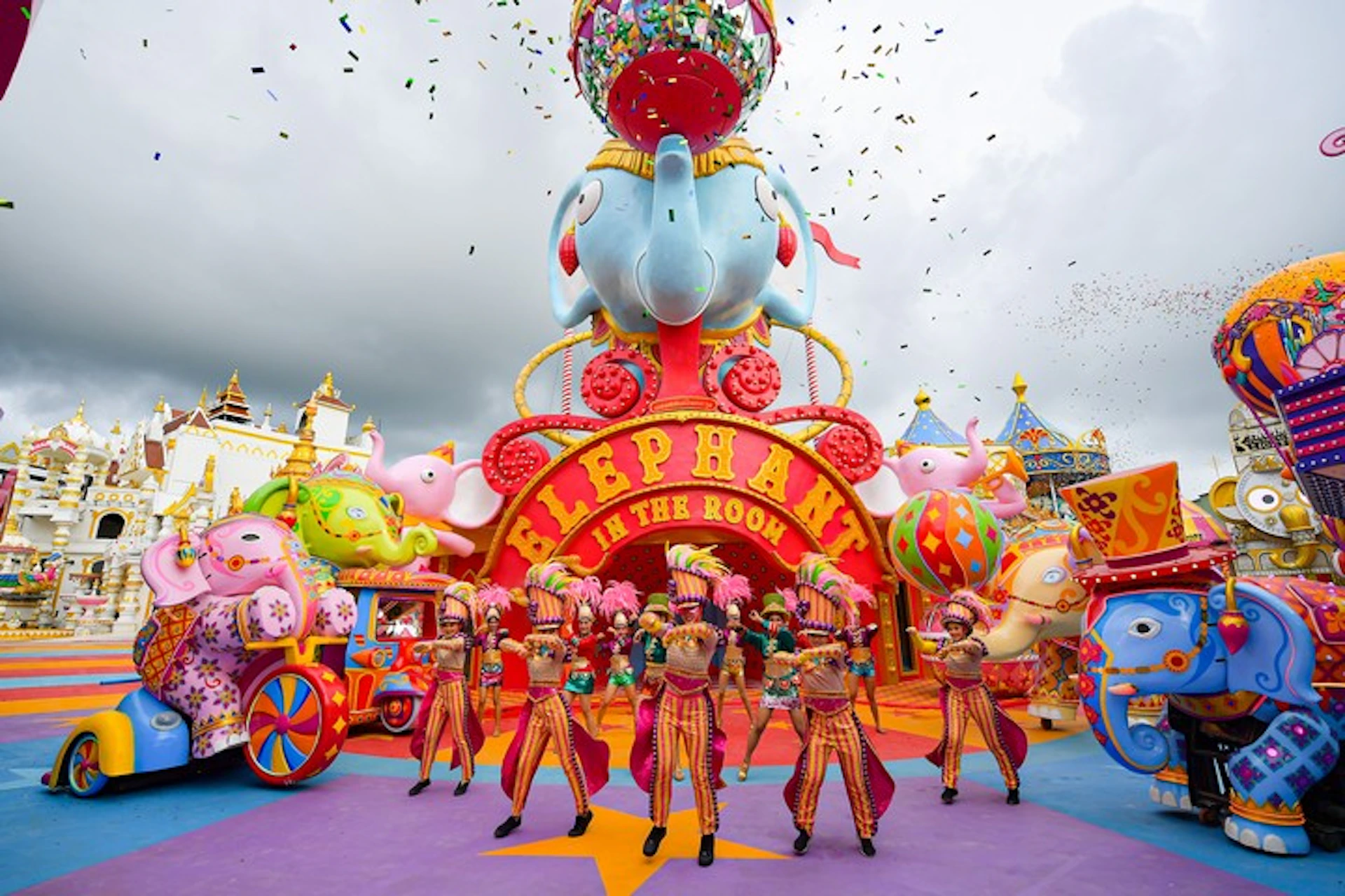 Performers in colorful costumes dance in front of a vibrant "Elephant in the Room" carnival display. Confetti falls from above.
