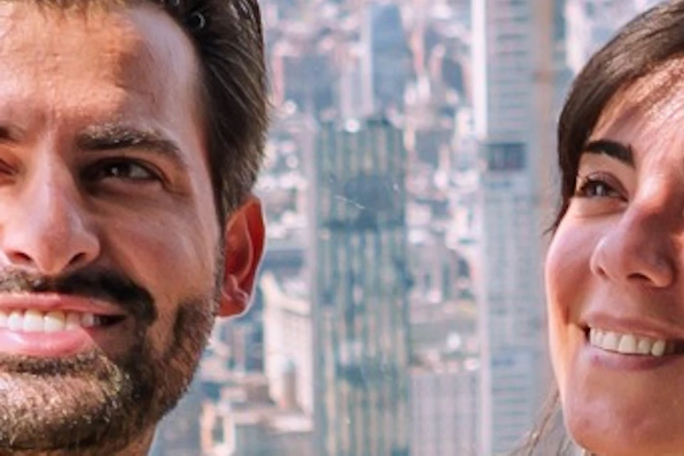 Two people taking a selfie on a glass observation deck with a city skyline, including the Empire State Building, in the background.