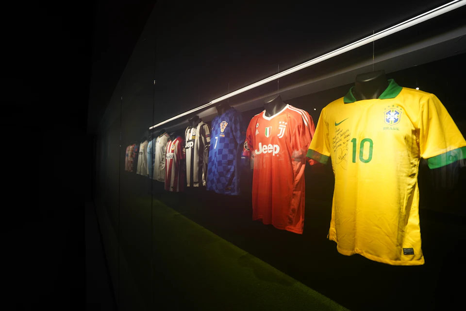 A lineup of soccer jerseys displayed in a glass case, including team uniforms in various designs and colors.