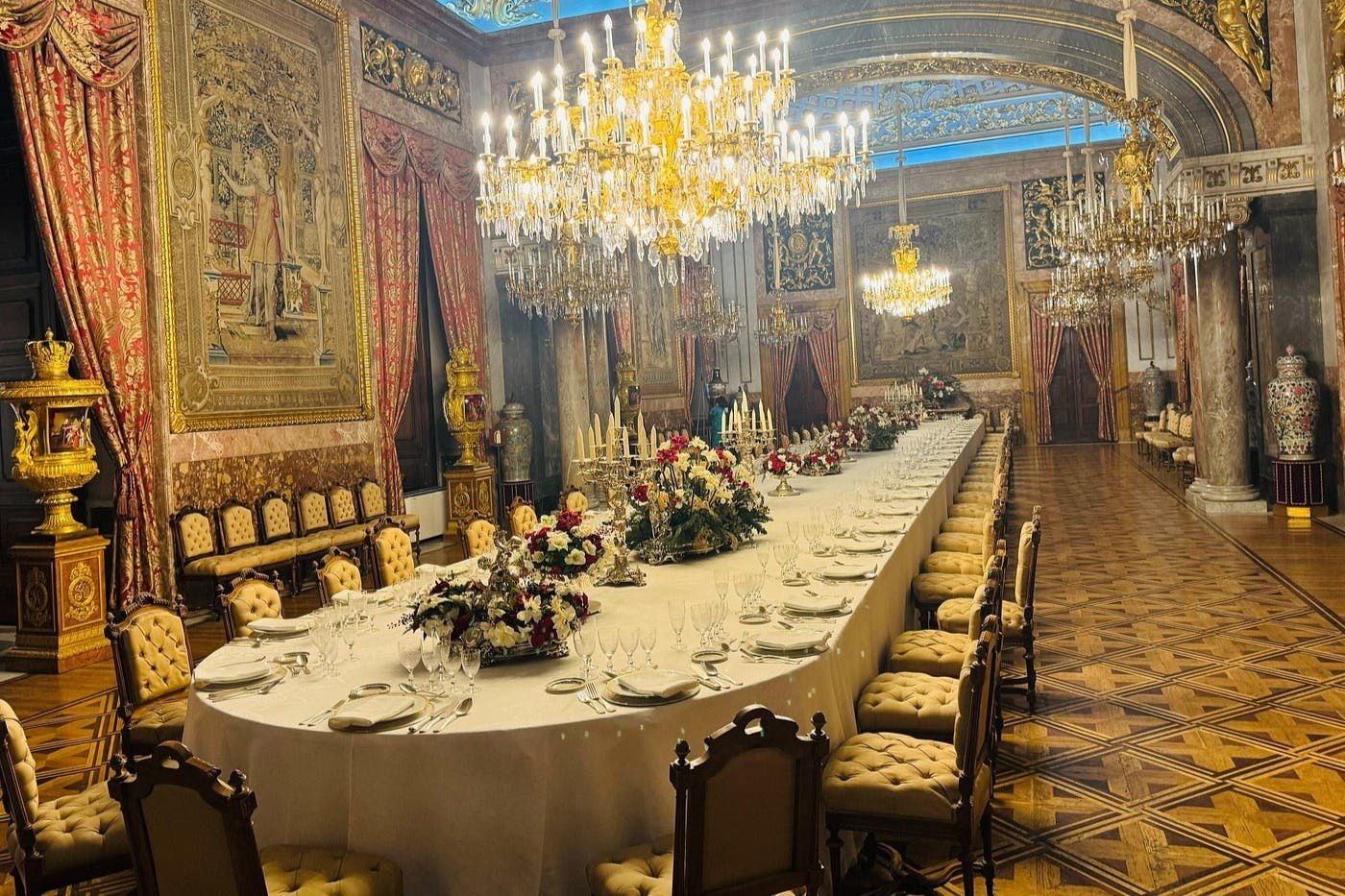 A luxurious dining hall with a long table set for a banquet, adorned with chandeliers, floral centerpieces, and ornate decor.