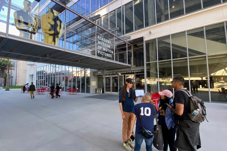 Four people gather outside the entrance of the Academy Museum of Motion Pictures, with a large Oscar statue visible behind glass above.