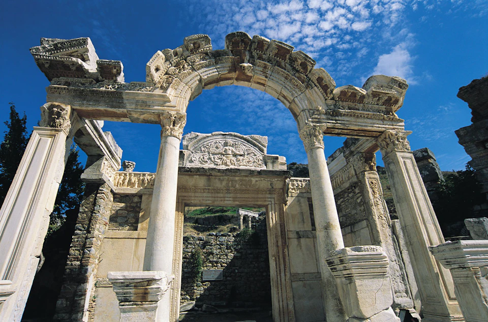 Ancient stone archway with ornate carvings, flanked by tall columns, against a partly cloudy blue sky.