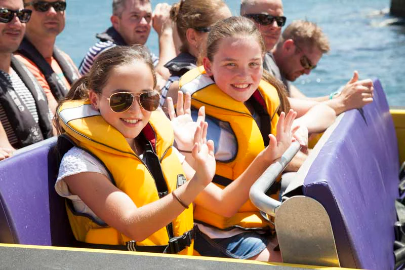 Two smiling girls wearing yellow life jackets and waving, surrounded by adults on a boat.
