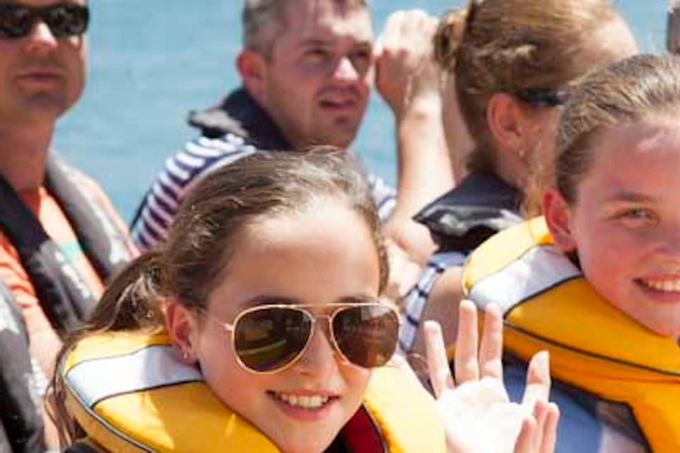 Two smiling girls wearing yellow life jackets and waving, surrounded by adults on a boat.