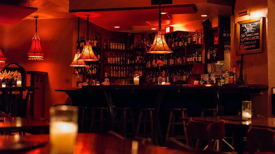 A dimly lit bar with bottles lined up on shelves, warm lighting from lamps, and wooden barstools in front of the counter.