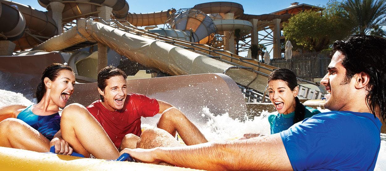 파란색 셔츠를 입은 두 사람이 워터 슬라이드를 타고 웃고 있으며, 배경에는 다단계 슬라이드 구조가 있습니다.