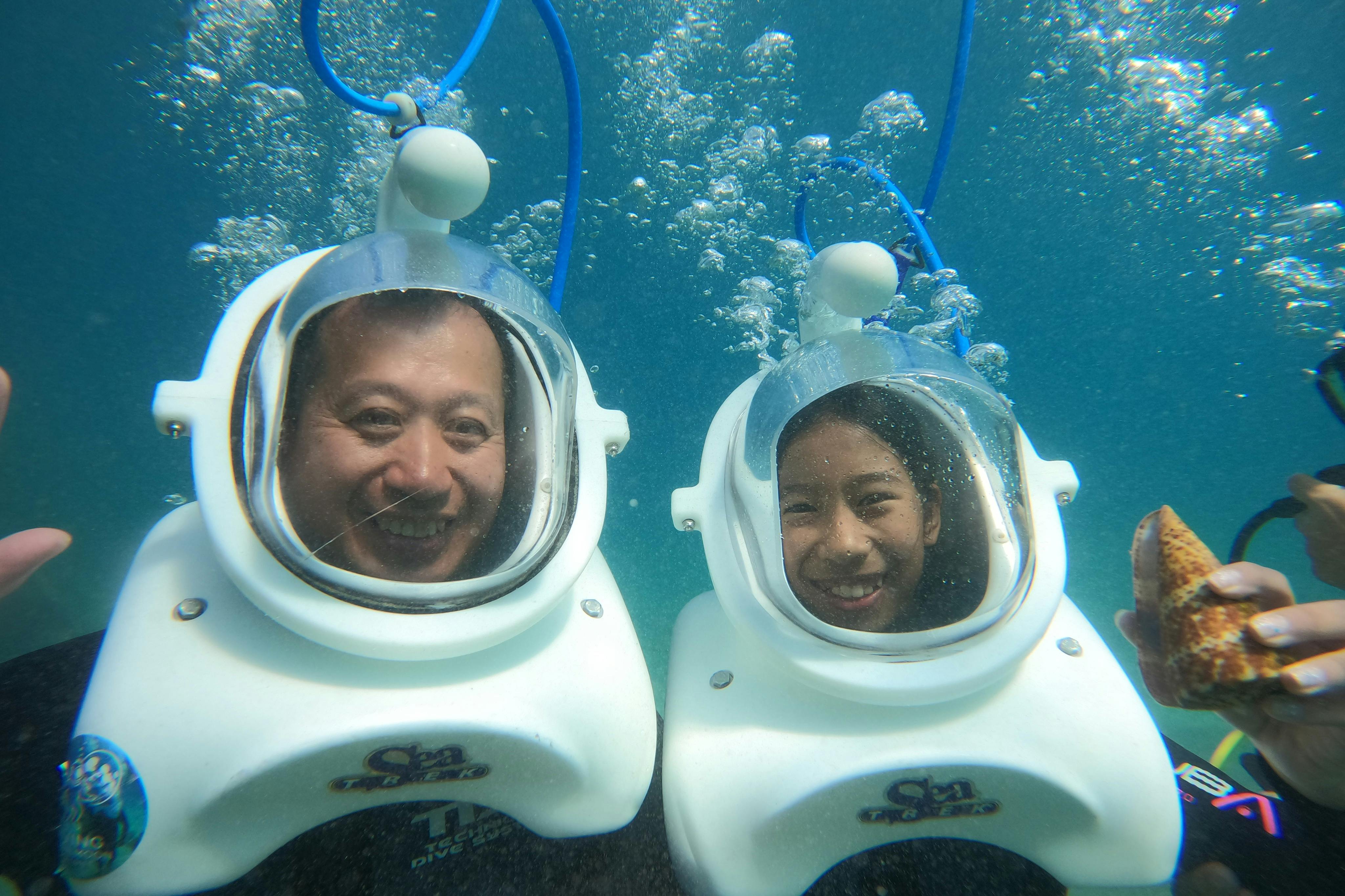Two people underwater wearing helmets with breathing tubes, smiling and surrounded by bubbling water.