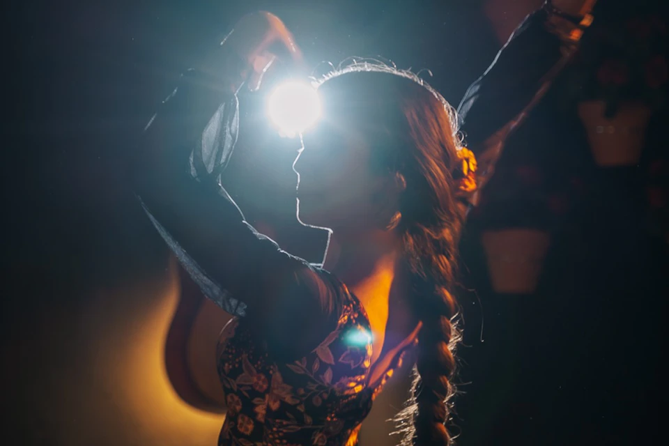 A person with a braided hairstyle raises an arm, illuminated by a bright light shining through their hand, creating a dramatic silhouette.