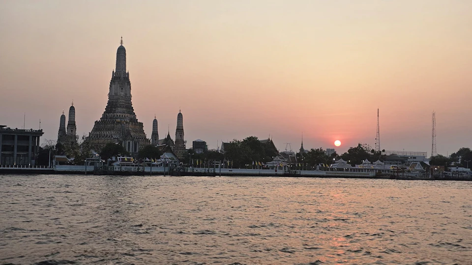Sunset view of Wat Arun temple by a calm river, with the sun low on the horizon, illuminating the sky with warm colors.