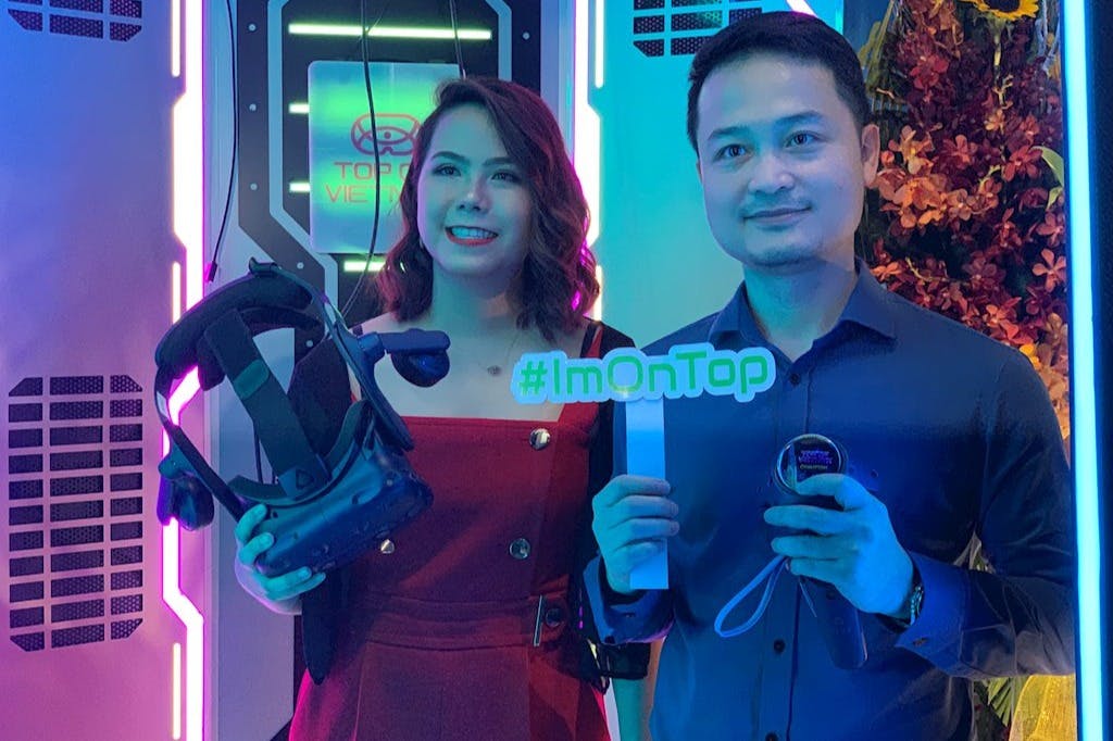 Two people smiling while holding VR equipment and a sign that says "#ImOnTop." There is a VR setup in the background.
