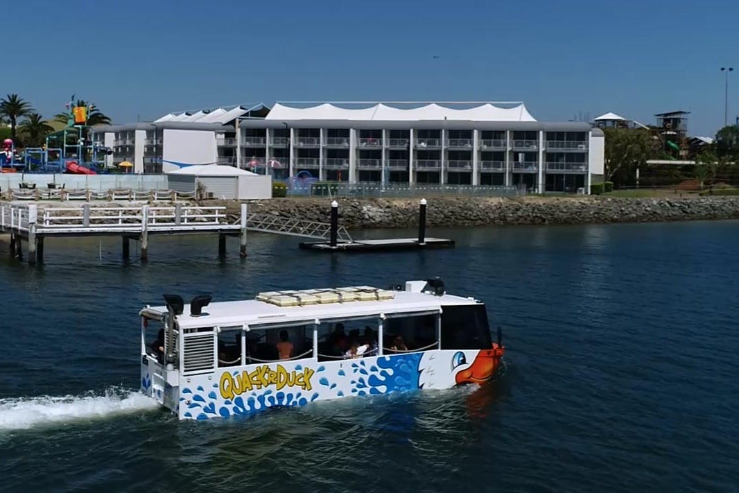 Prohlídkový autobus, který má vypadat jako loď, se jmenuje Quack'R' Duck a pohybuje se po vodě v blízkosti doku s budovami v pozadí.