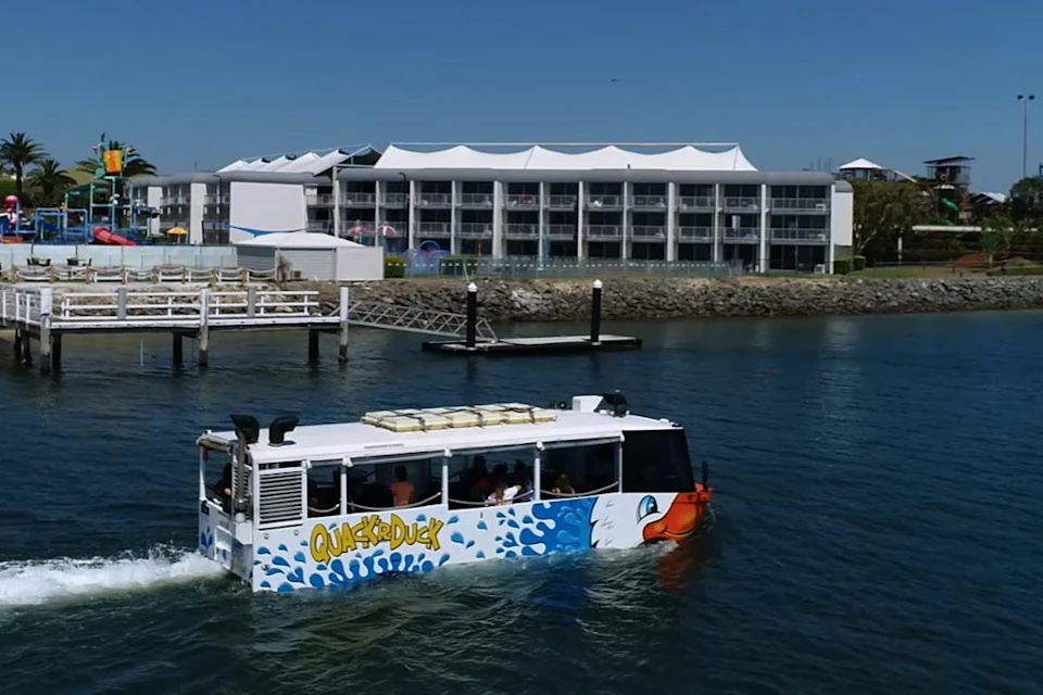 A tour bus designed to look like a boat, named Quack'R'Duck, moves through water near a dock with buildings in the background.
