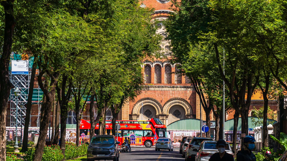 A street lined with trees, cars parked on the sides, and a red double-decker bus passing in front of a brick building.