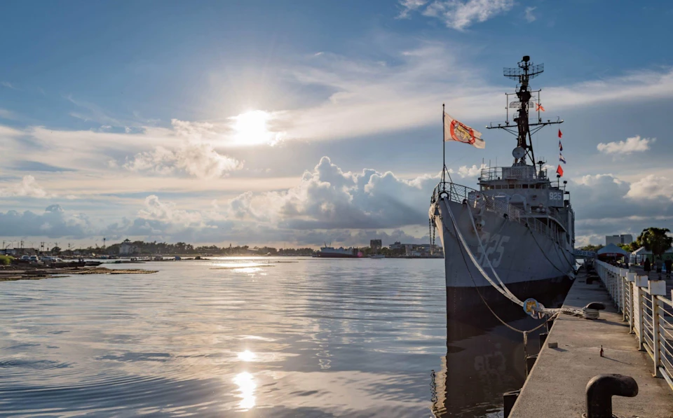 A large ship docked by a calm waterfront under a partly cloudy sky with the sun shining. A flag is flying on the ship's mast.