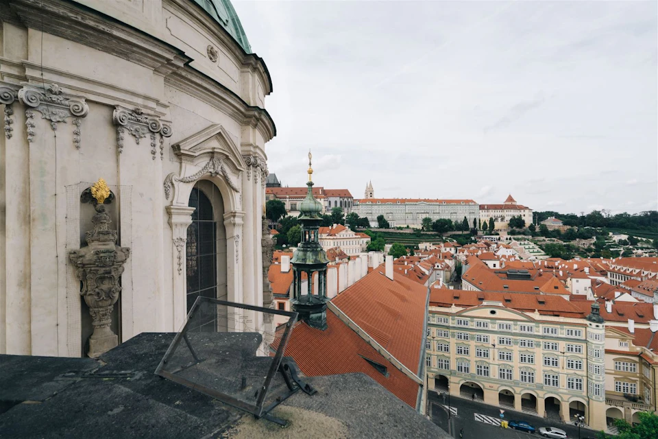A view of historic buildings with red rooftops and ornate architecture under a cloudy sky. A glass panel and railing are visible in the foreground.