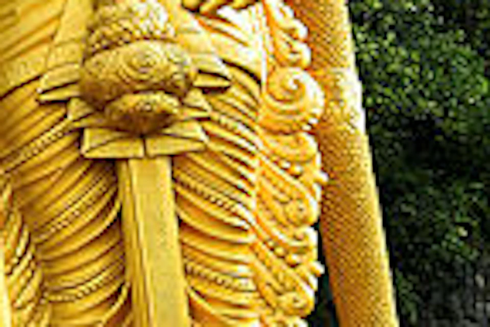 Large golden statue of a deity stands near a colorful, crowded staircase leading to a cave entrance amidst lush greenery.