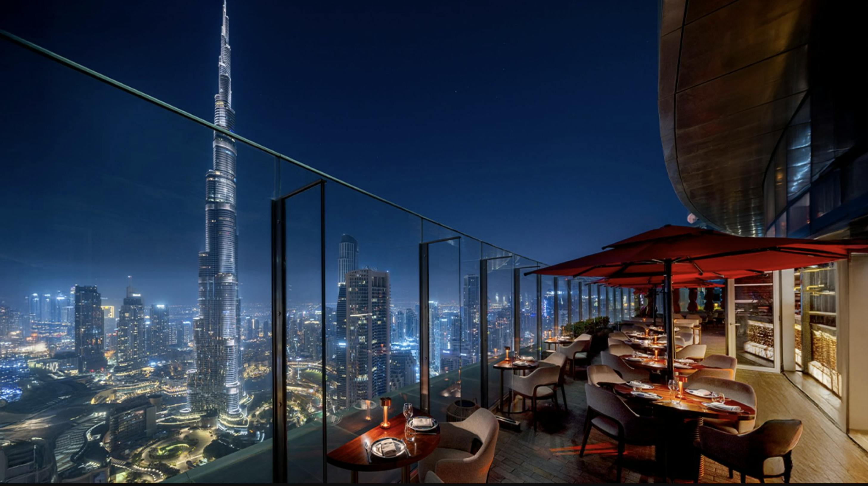 Un restaurant al terrat amb paraigües vermells i taules amb vistes a l'horari de la ciutat amb un gran gratacel il·luminat a la nit.